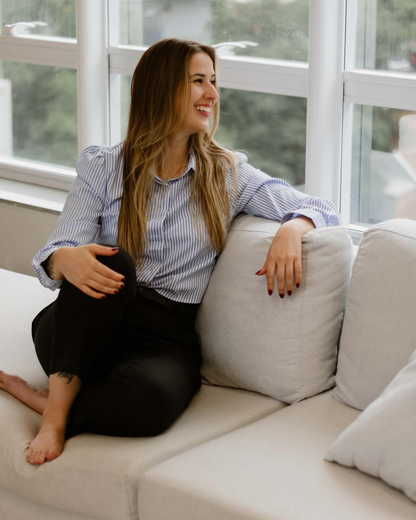 Amanda Rocca sorrindo, sentada em um sofá próximo à janela